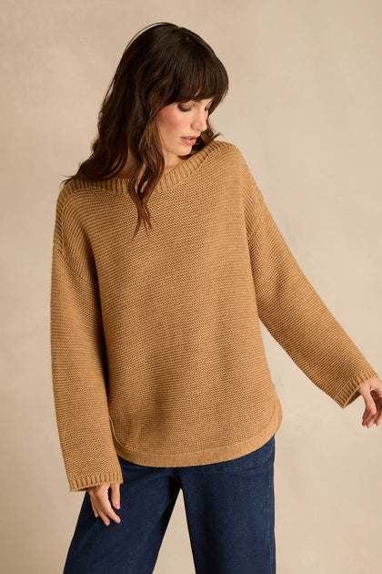 A person wearing the Camel Ribbed Wool Blend Button Jumper stands against a beige background, looking down and to the side. The ribbed wool blend jumper pairs effortlessly with dark blue pants for an understated, classic look.