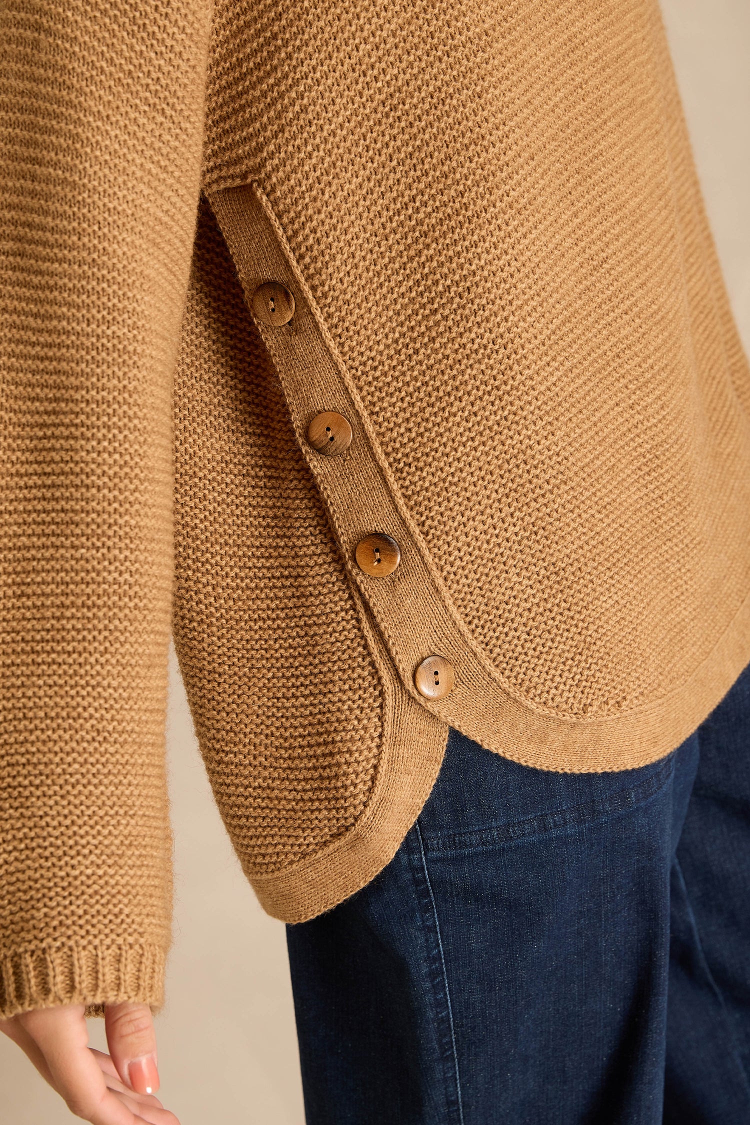Close-up of a person wearing the Camel Ribbed Wool Blend Button Jumper with four side buttons and dark blue jeans, hand slightly extended.