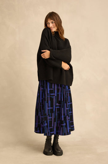 A woman stands against a neutral background, looking to the side and smiling, dressed in a Black Merino Ribbed Turtle Neck Jumper, a blue and black patterned midi skirt, and black boots.