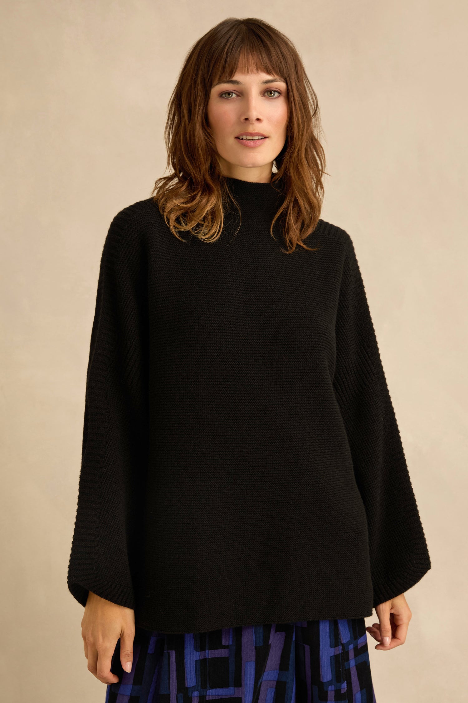 A woman wearing the Black Merino Ribbed Turtle Neck Jumper and a blue-patterned skirt stands against a plain beige background.