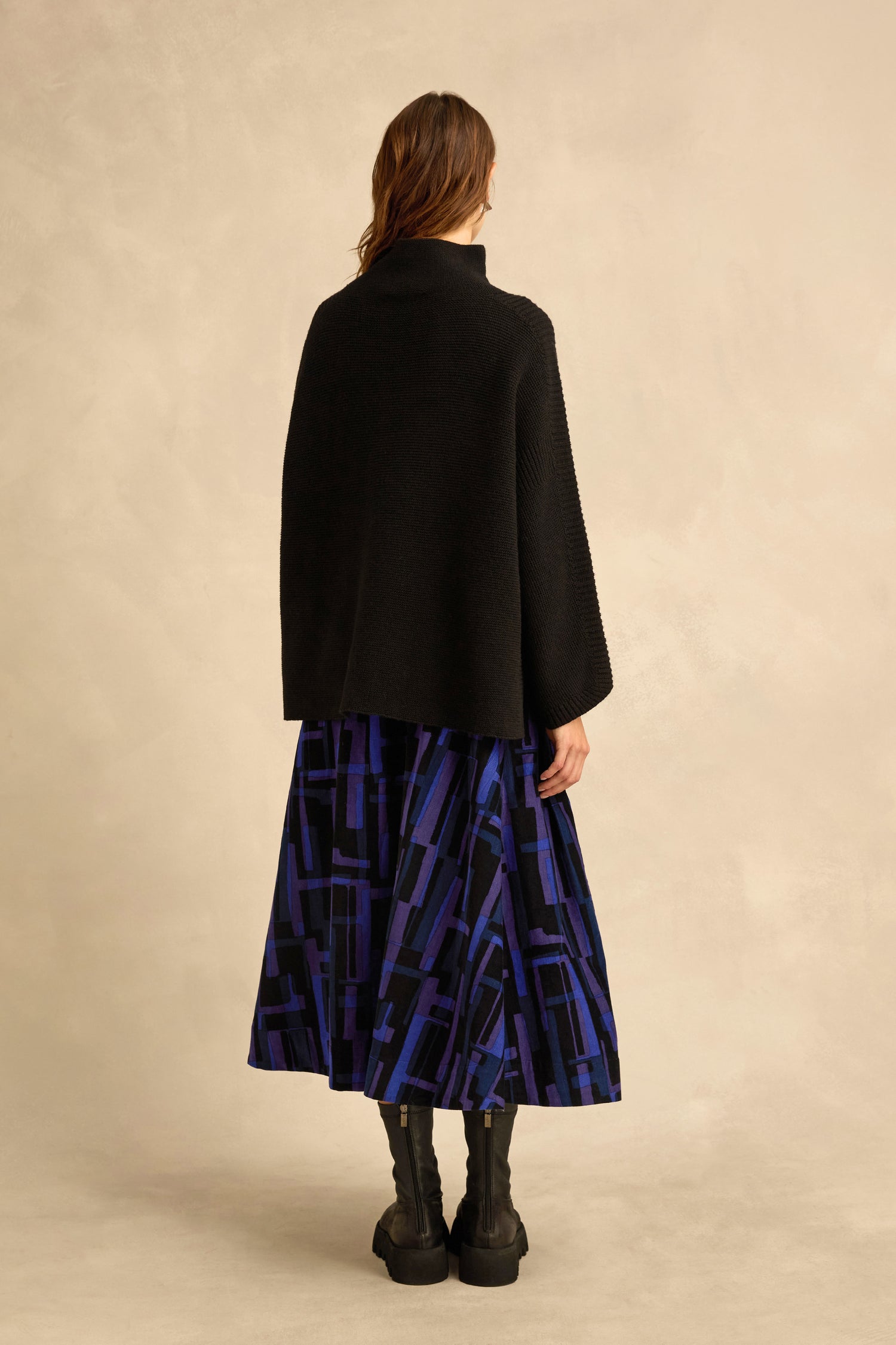 A person stands facing away, wearing the Black Merino Ribbed Turtle Neck Jumper, a blue and black geometric-patterned skirt, and black boots against a neutral background.