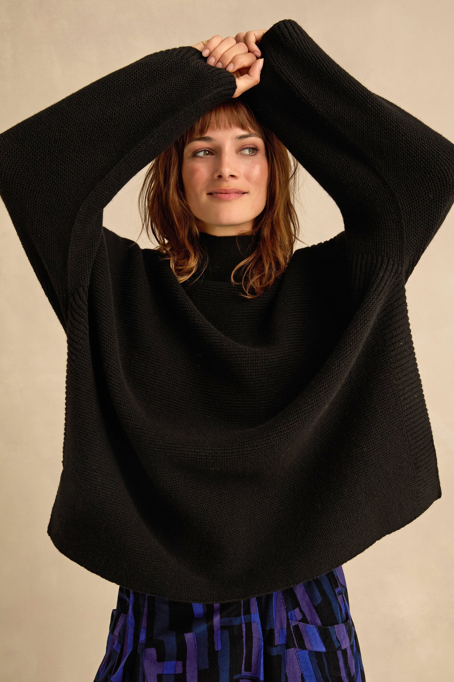A woman wearing a black Merino ribbed turtle neck jumper and a blue patterned skirt stands with her arms raised above her head, looking to the side against a neutral background.