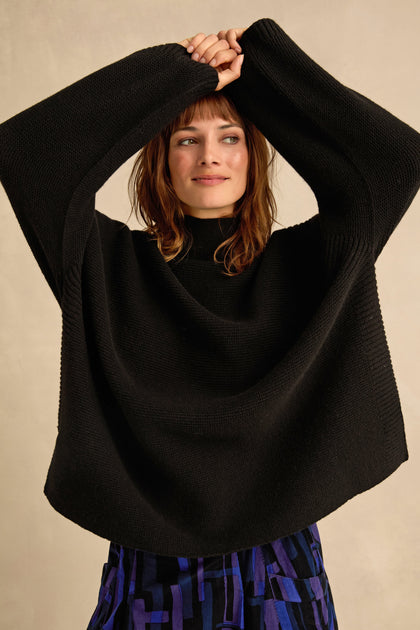 A woman wearing a black Merino ribbed turtle neck jumper and a blue patterned skirt stands with her arms raised above her head, looking to the side against a neutral background.