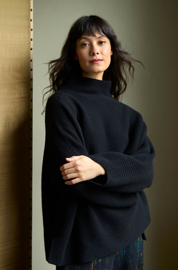 A person wearing a black, long-sleeved Black Merino Ribbed Turtle Neck Jumper with their arms crossed.