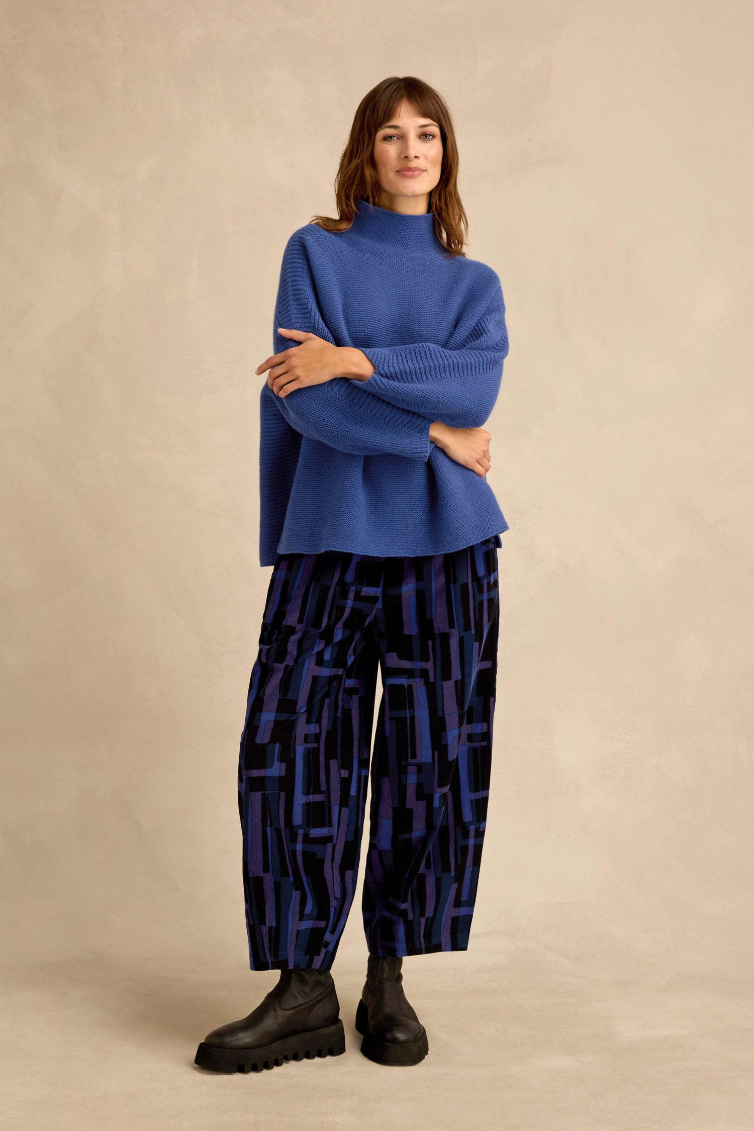 A woman stands against a beige backdrop, wearing the Royal Blue Merino Ribbed Turtle Neck Jumper, paired with patterned wide-leg pants in dark blue and black and black chunky shoes. Her arms are crossed as she looks forward.