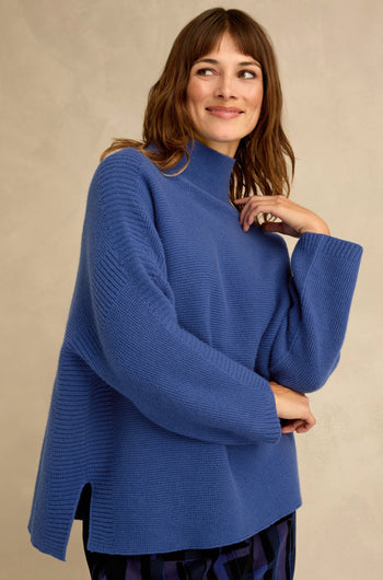 A woman wearing the Royal Blue Merino Ribbed Turtle Neck Jumper, featuring a relaxed fit with side slits and a high neckline, stands against a plain light beige background, smiling and gazing to the side.