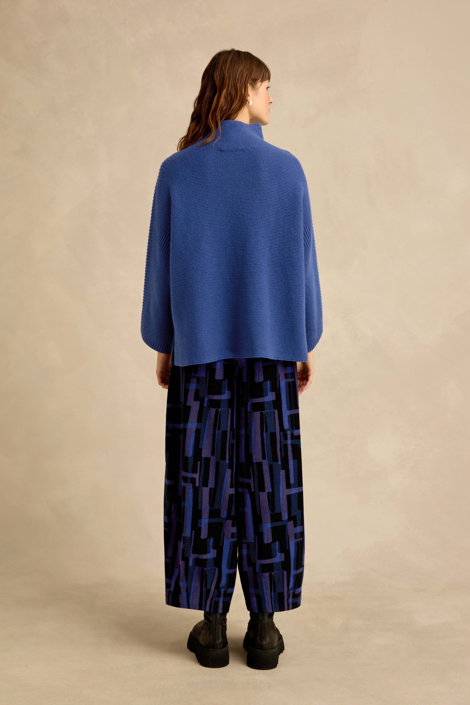 A woman stands facing away, wearing the Royal Blue Merino Ribbed Turtle Neck Jumper, patterned wide-leg pants, and black shoes against a beige backdrop.
