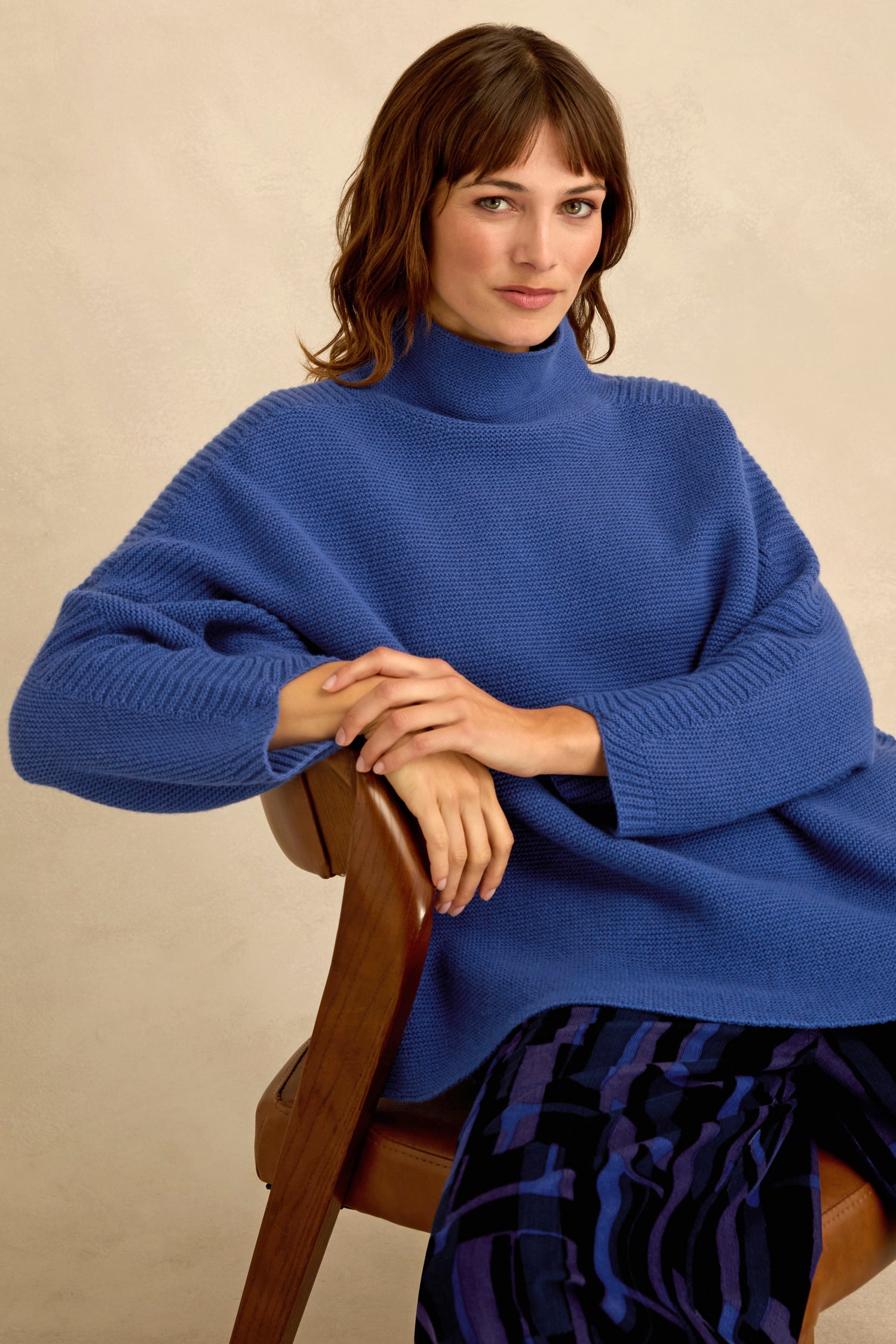 Woman with brown hair in a Royal Blue Merino Ribbed Turtle Neck Jumper and patterned pants sits on a wooden chair, facing forward with her hands resting on her knee.