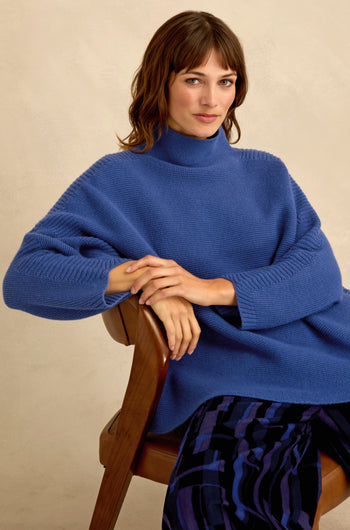Woman with brown hair in a Royal Blue Merino Ribbed Turtle Neck Jumper and patterned pants sits on a wooden chair, facing forward with her hands resting on her knee.