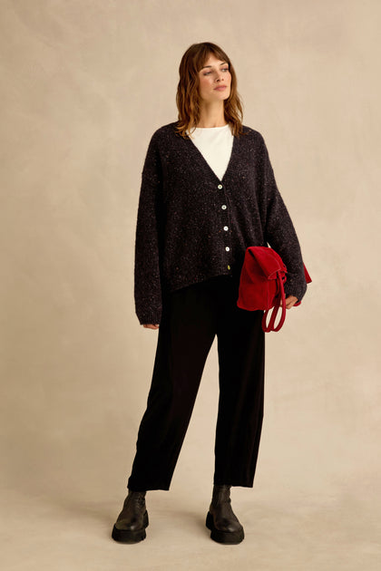 A woman stands against a beige background, wearing the Night Alpaca Blend Cropped Cardi, a white shirt, black pants, black boots, and holding a red bag in her left hand.