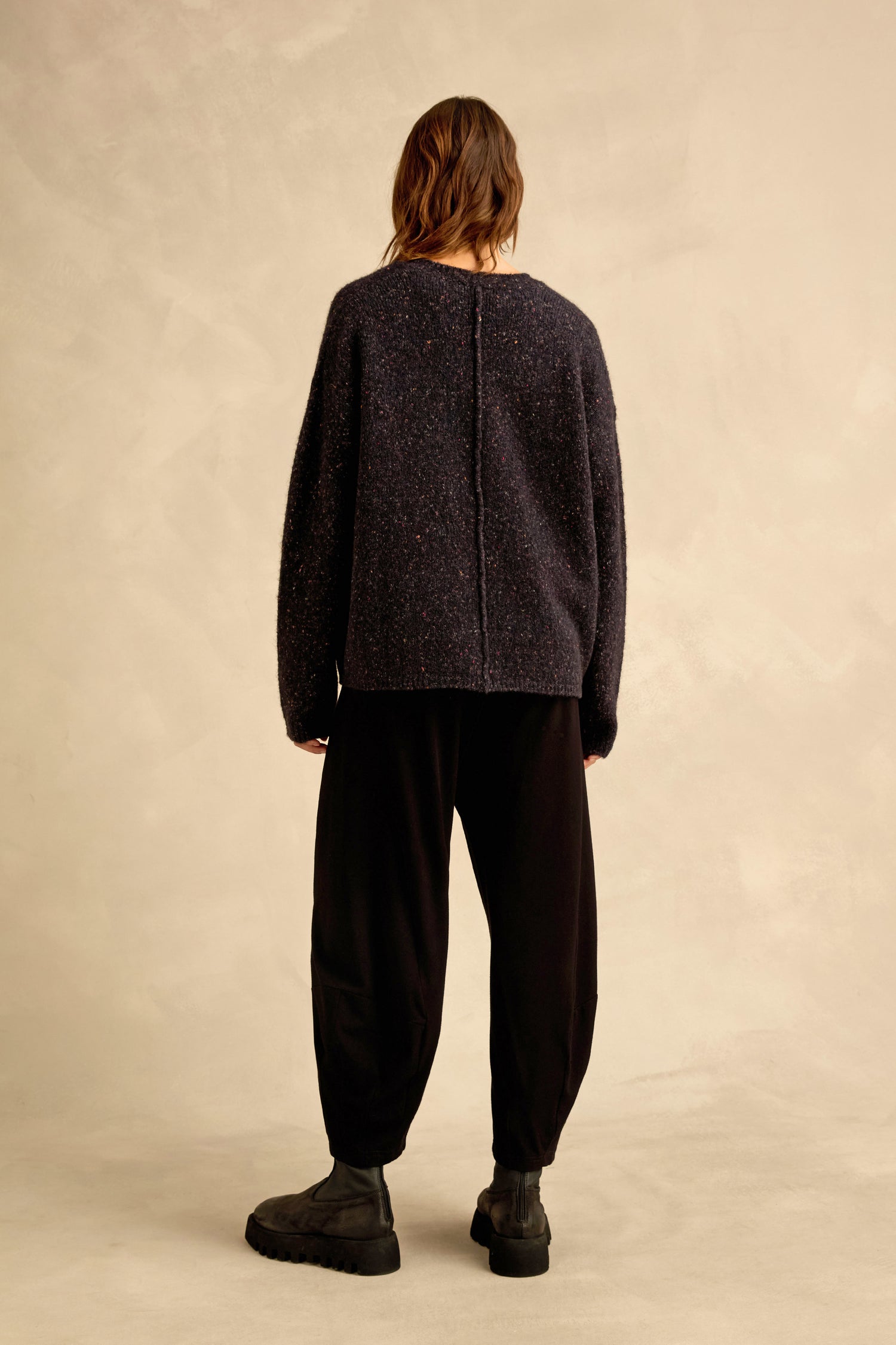 A person stands with their back to the camera, wearing the Night Alpaca Blend Cropped Cardi, loose black pants, and black boots against a beige background.