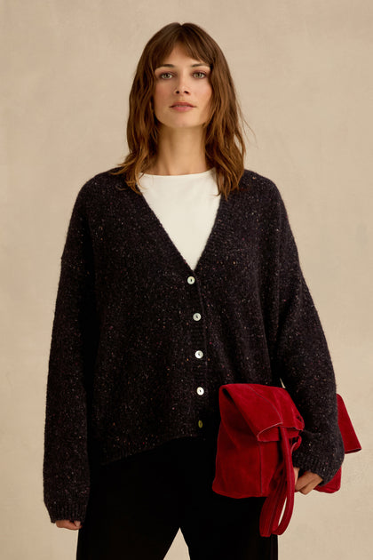 A woman stands against a plain background, wearing the Night Alpaca Blend Cropped Cardi in black over a white shirt and holding a large red suede bag.