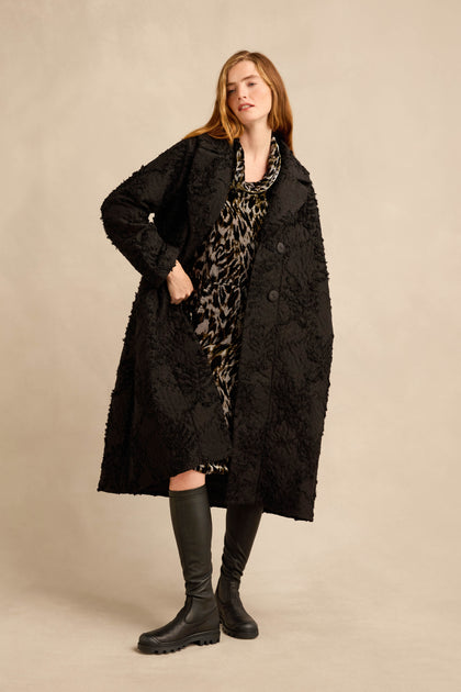A woman wearing the Textural Big Collar Coat in black over a leopard print dress and black knee-high boots, standing against a beige background.