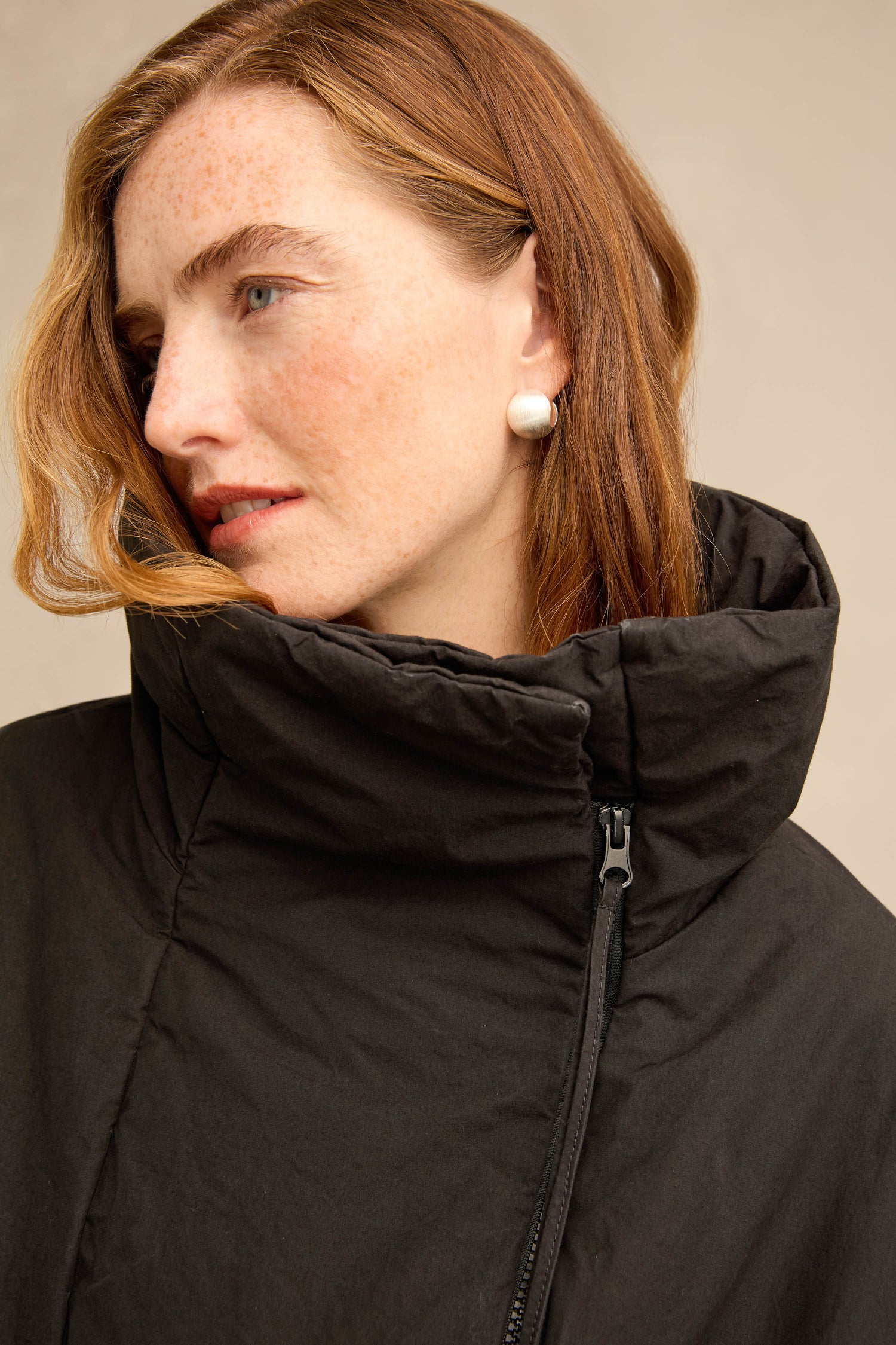 A woman with red hair and freckles wears the Big Pocket Puffer Jacket, a black oversized puffer with a high collar and zip fastening, paired with round silver stud earrings.