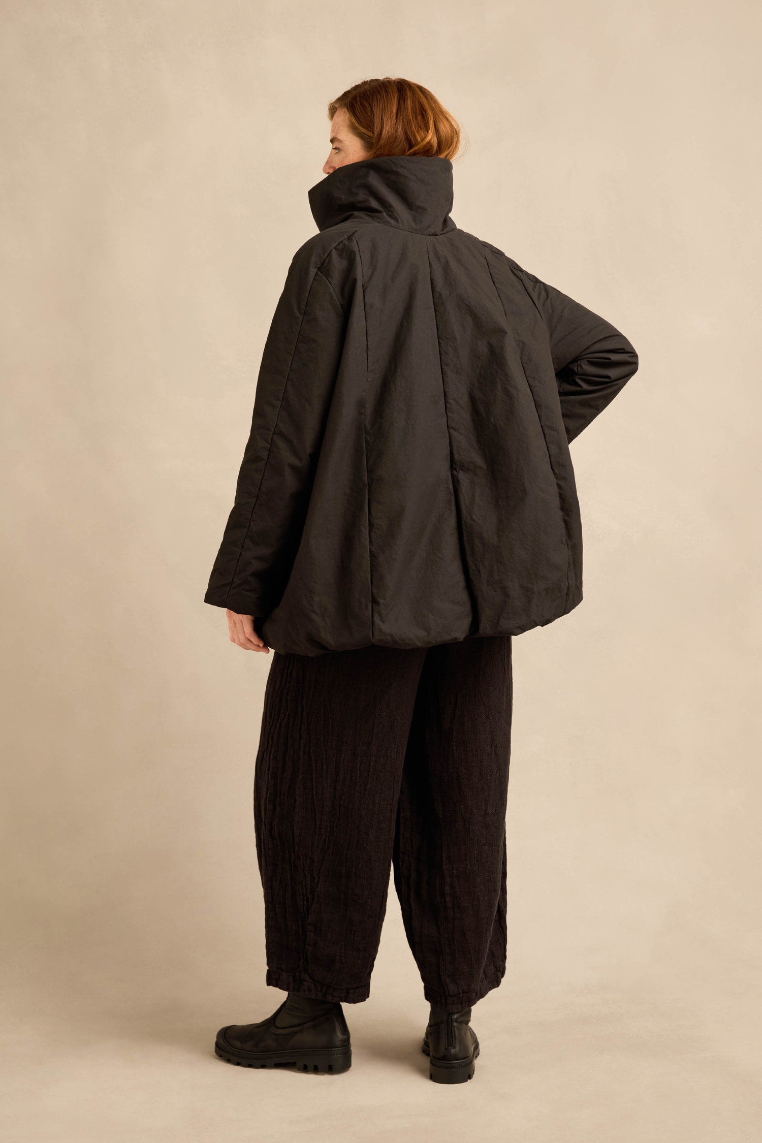 A person stands with their back turned, wearing the Big Pocket Puffer Jacket, black textured pants, and black boots against a plain beige background.
