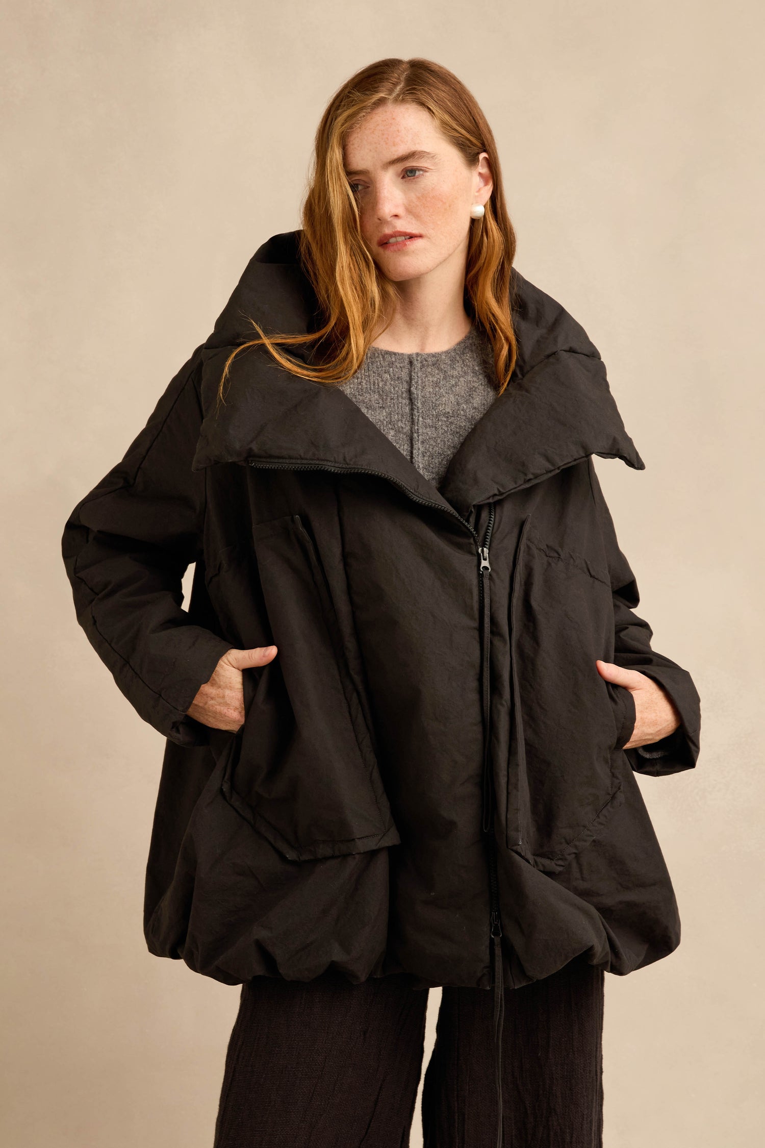 A person wearing the Big Pocket Puffer Jacket—a loose-fitting black cocoon silhouette coat with a wide collar and side pockets—stands against a plain beige background.