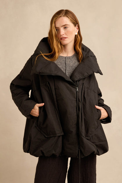 A person wearing the Big Pocket Puffer Jacket—a loose-fitting black cocoon silhouette coat with a wide collar and side pockets—stands against a plain beige background.