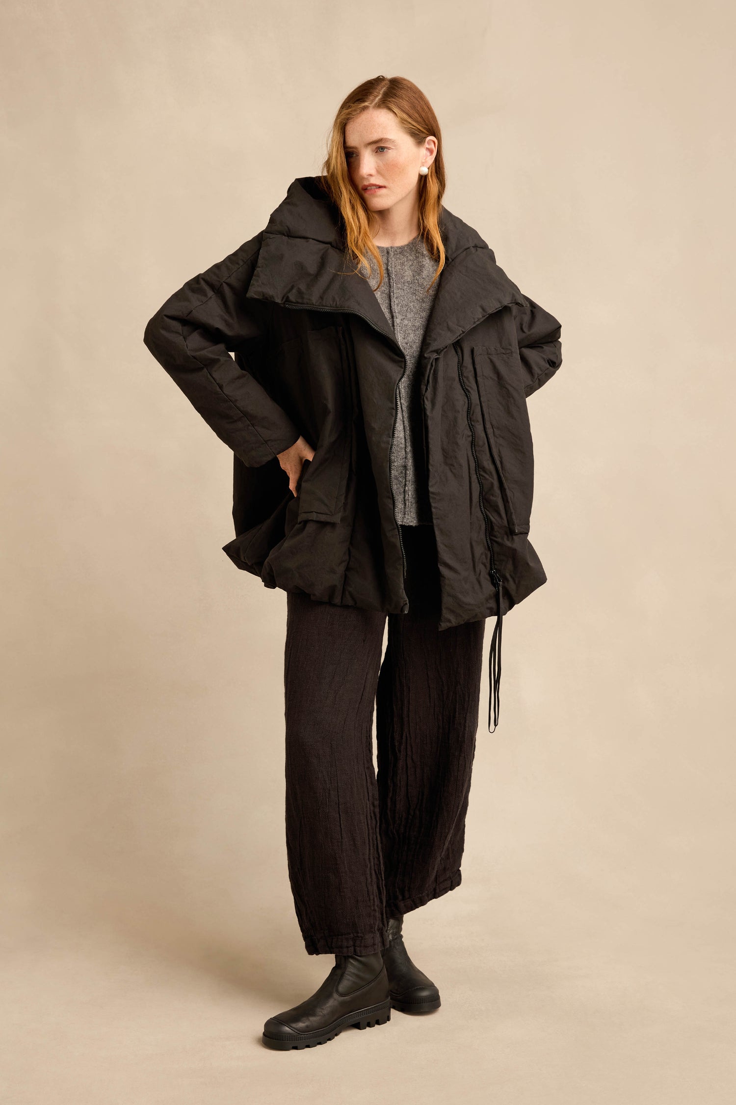 A person stands against a plain beige background, wearing the Big Pocket Puffer Jacket in black styled with a gray top, black wide-leg pants, and black boots.