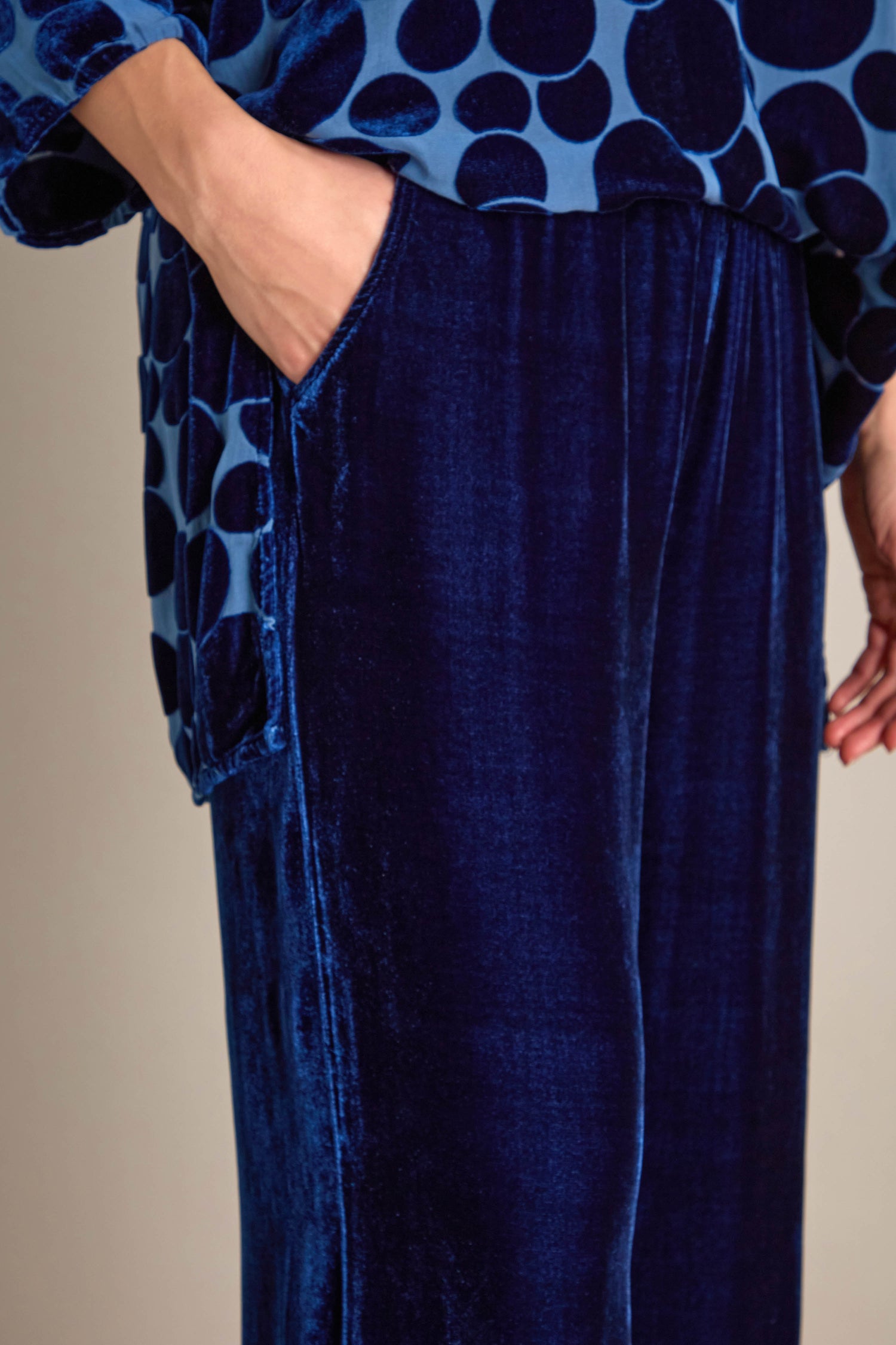 A person dressed in a blue velvet ensemble is wearing a polka dot top paired with Silk Velvet Trousers that have a plain design and cropped length. One hand rests casually in their pants pocket, while the elasticated waistband provides both style and comfort.