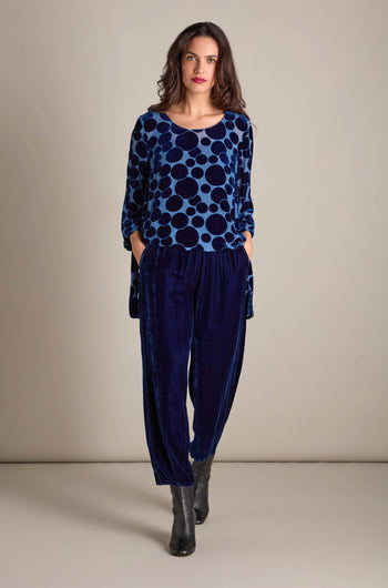 A woman wearing a blue polka dot top and the Silk Velvet Trousers, featuring an elasticated waistband, stands with her hands in the pockets against a neutral background.