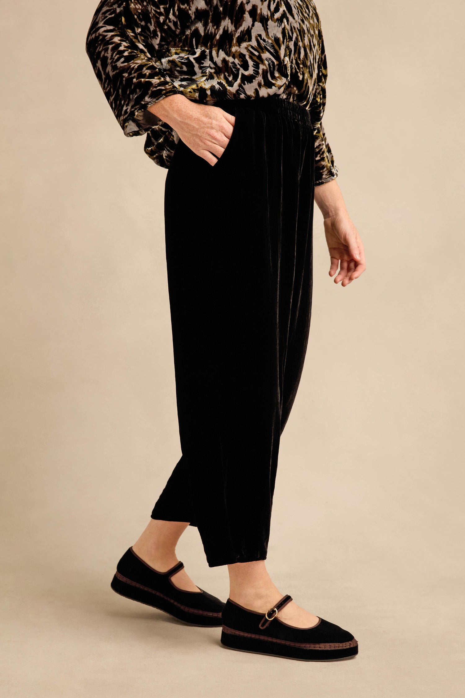 Person standing sideways wearing a patterned long-sleeve top, the Black Silk Velvet Trouser with a relaxed fit and tapered leg, and black Mary Jane shoes against a plain beige background.