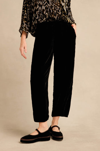 A person wearing the Black Silk Velvet Trouser, featuring a relaxed fit and tapered leg, paired with a patterned long-sleeve top and black Mary Jane shoes, stands against a plain beige background.
