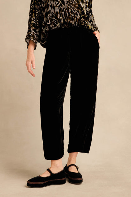 A person wearing the Black Silk Velvet Trouser, featuring a relaxed fit and tapered leg, paired with a patterned long-sleeve top and black Mary Jane shoes, stands against a plain beige background.