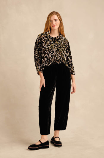 A person stands against a beige backdrop wearing a loose animal-print top, the Black Silk Velvet Trouser with a tapered leg and relaxed fit, and black platform shoes.