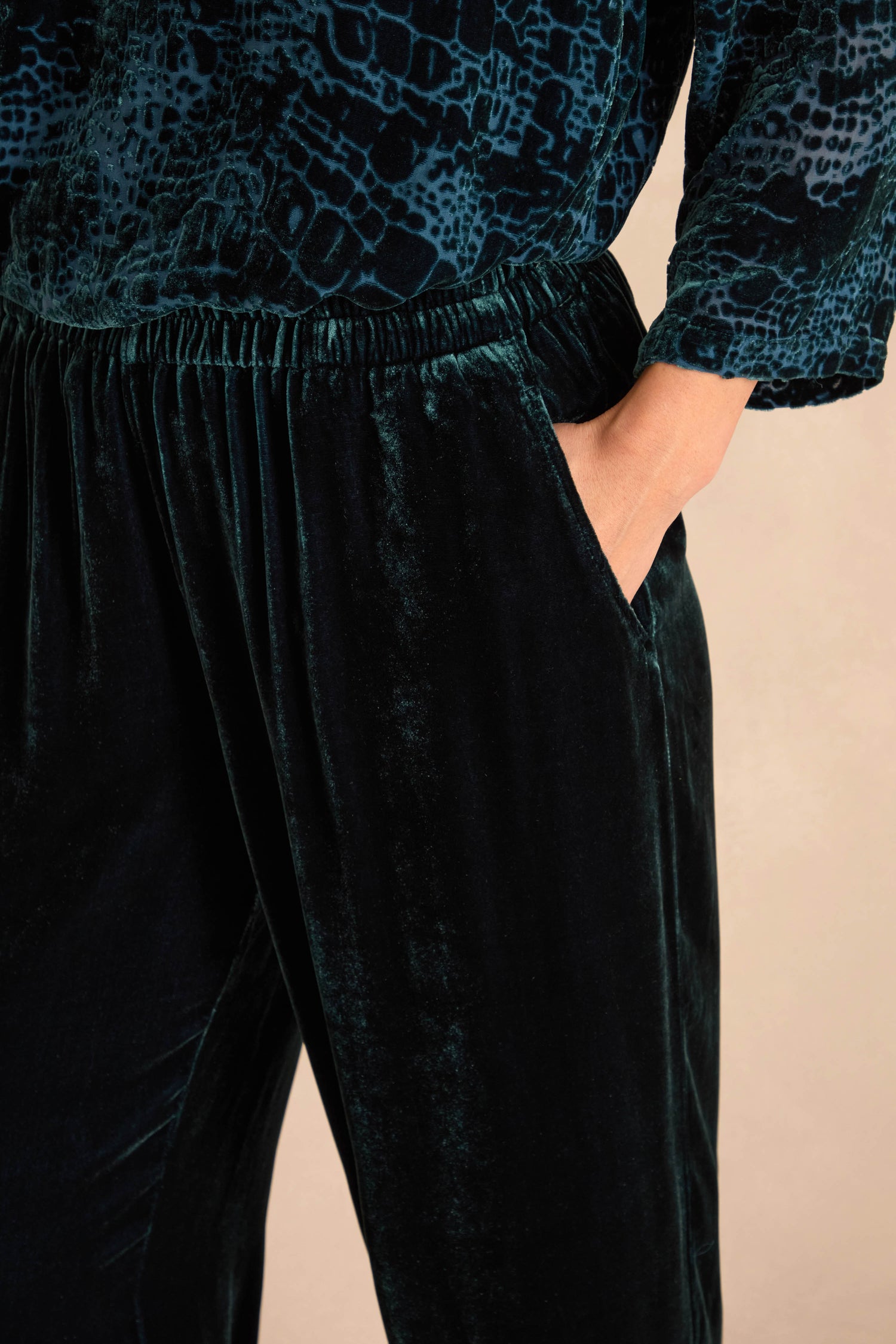 Person wearing Deep Turquoise Silk Velvet Trousers with an elasticated waist, hand in pocket, and a textured dark blue patterned top.