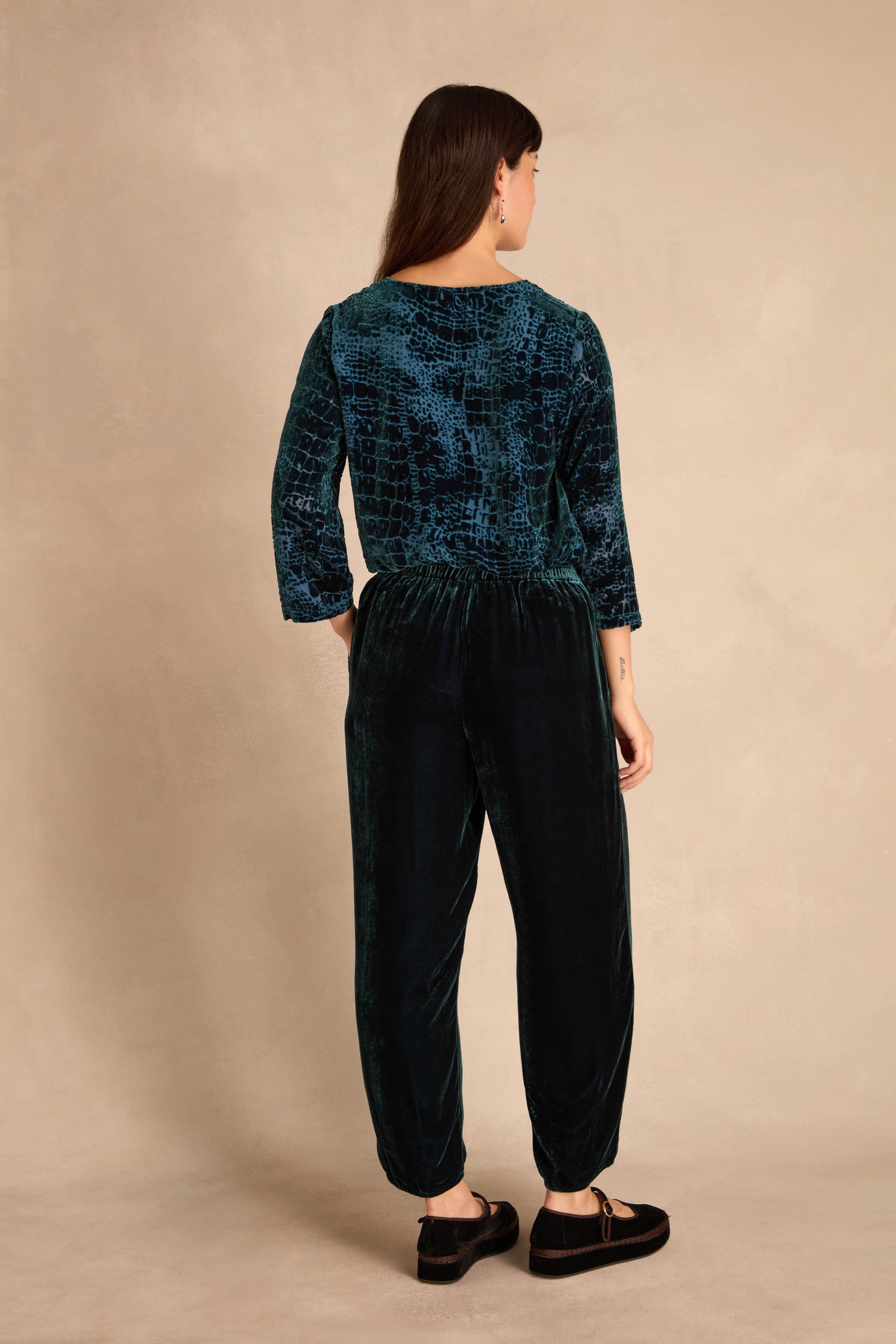 A woman faces away, wearing Deep Turquoise Silk Velvet Trousers with a dark teal textured top and black shoes against a beige background.