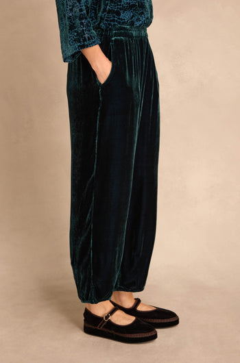 Person wears Deep Turquoise Silk Velvet Trousers with elasticated waist, hands in pockets, and black Mary Jane shoes; shown waist down against a neutral background.
