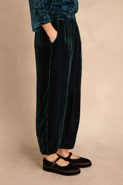 Person wears Deep Turquoise Silk Velvet Trousers with elasticated waist, hands in pockets, and black Mary Jane shoes; shown waist down against a neutral background.