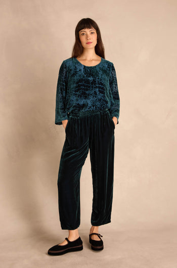 Woman stands against a beige wall wearing a loose dark teal patterned top and Deep Turquoise Silk Velvet Trousers with an elasticated waist, hands in pockets, and black flats.