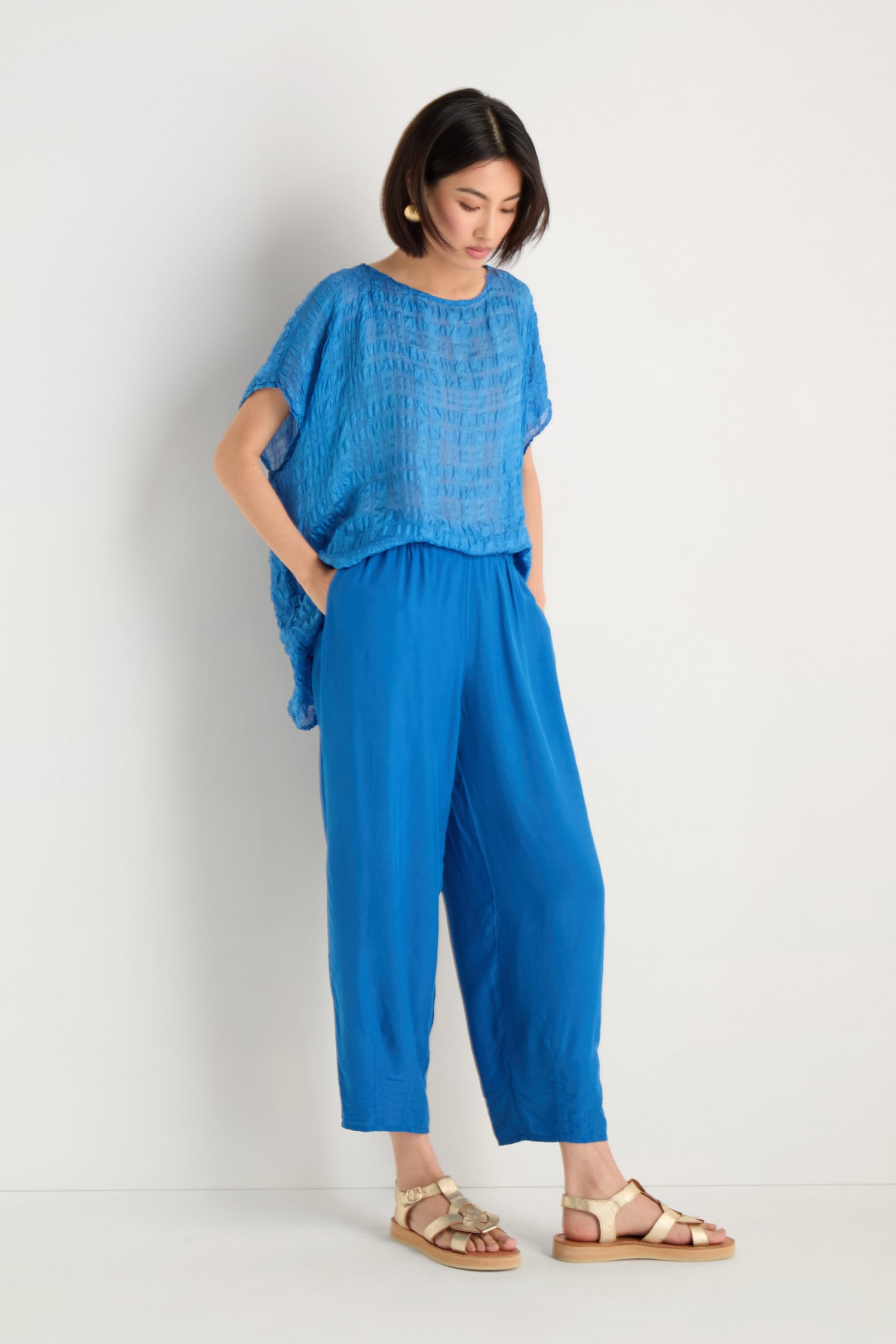 A person stands against a white background wearing Relaxed Bamboo Silk Trousers in blue with an elasticated waist, paired with a loose top and beige sandals, hands in pockets and head slightly tilted down.