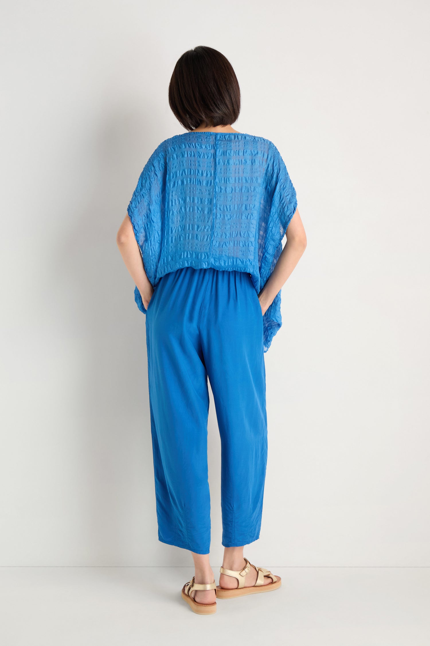 Facing away, a person wears a relaxed-fit blue top, Relaxed Bamboo Silk Trouser with an elasticated waist, and beige sandals against a light background.