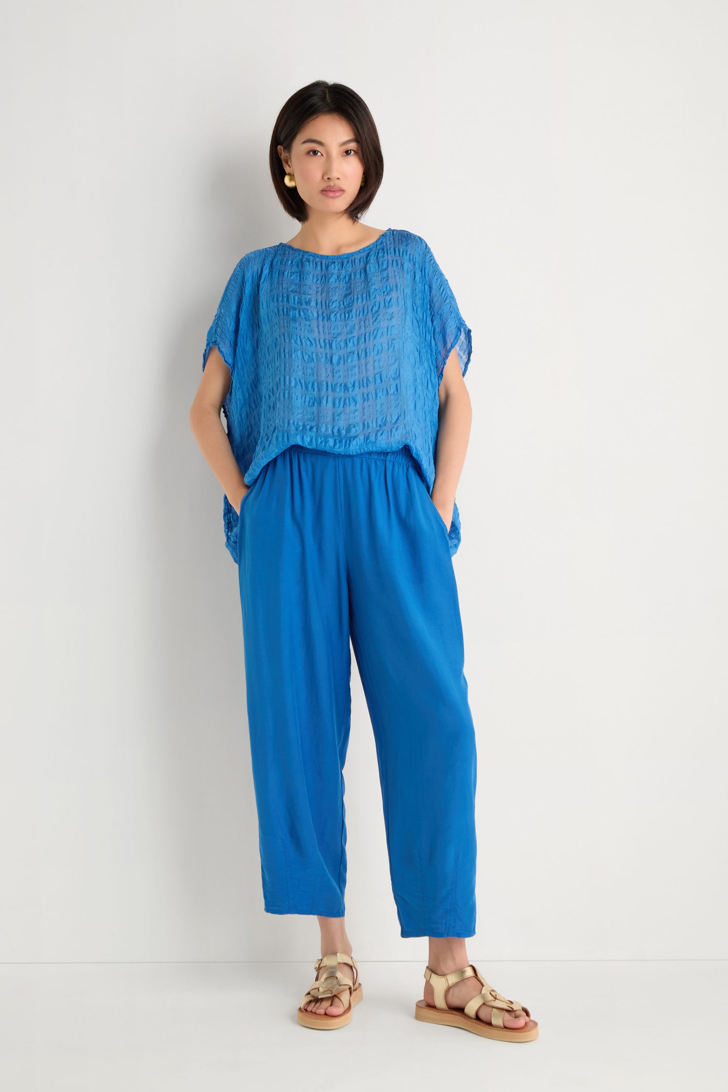 A woman stands against a white background, wearing a loose blue textured top, Relaxed Bamboo Silk Trouser with an elasticated waist, beige sandals, and gold earrings.