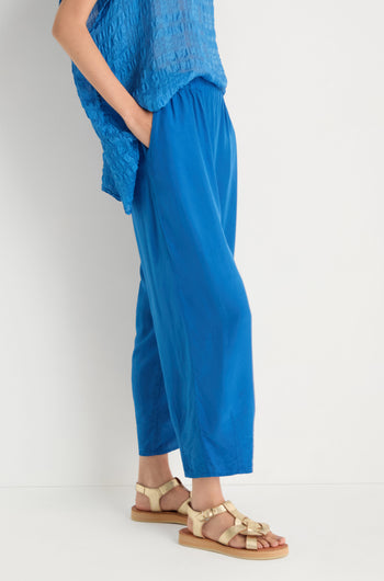 Model wears Relaxed Bamboo Silk Trouser in blue with a relaxed fit and elasticated waist, paired with a textured blue top and beige strappy sandals, posing against a white background with one hand in pocket.