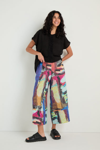 A woman stands against a white wall, wearing a loose black top, Abstract Print Wide Leg Trousers, and black sandals.