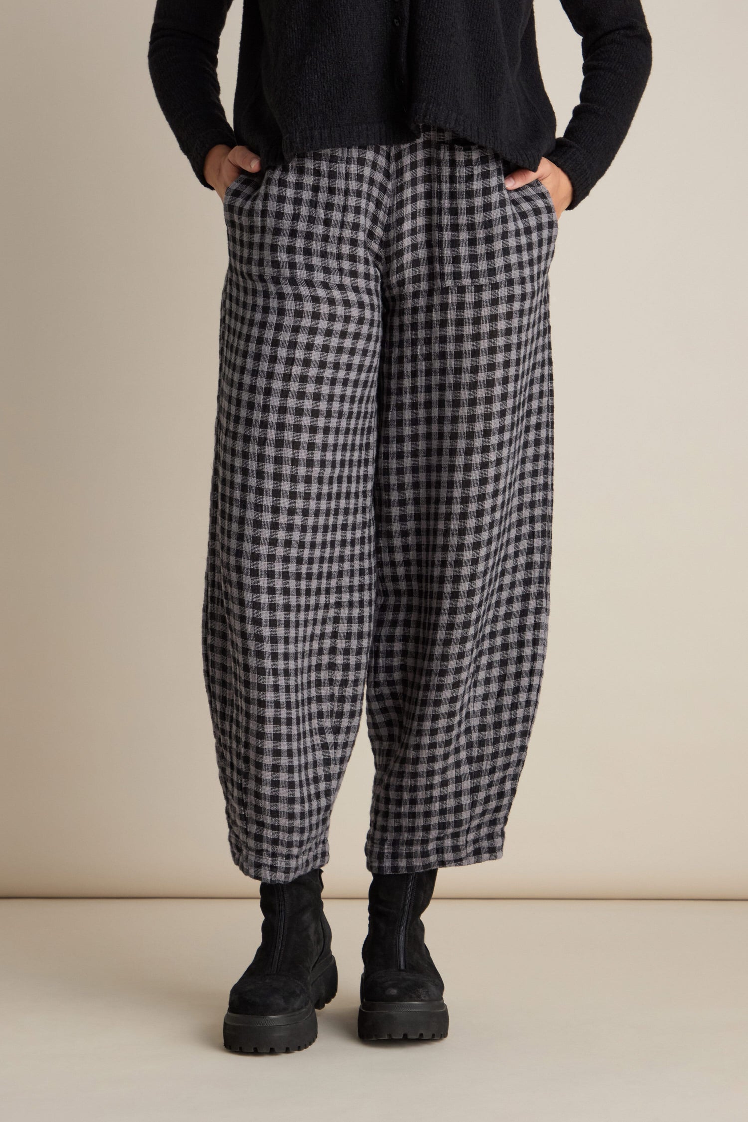 A person wearing the Linen Check Trousers featuring a black and grey pattern and an elasticated waist, paired with black boots and a black top, stands against a beige background with hands in pockets.