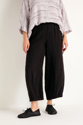Wearing a loose, light-colored top crafted from breathable natural fabric, paired with the Textured Linen Charcoal Bubble Trousers and black lace-up shoes, standing against a white wall.
