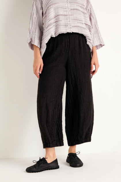 Wearing a loose, light-colored top crafted from breathable natural fabric, paired with the Textured Linen Charcoal Bubble Trousers and black lace-up shoes, standing against a white wall.