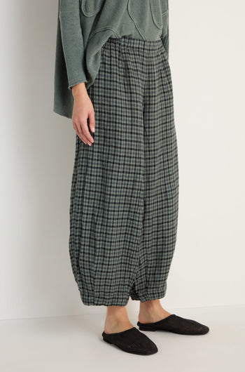 Wearing Check Linen Bubble Trousers in green and grey with a loose silhouette, paired with an olive green top and black slip-on shoes, standing against a plain light background.