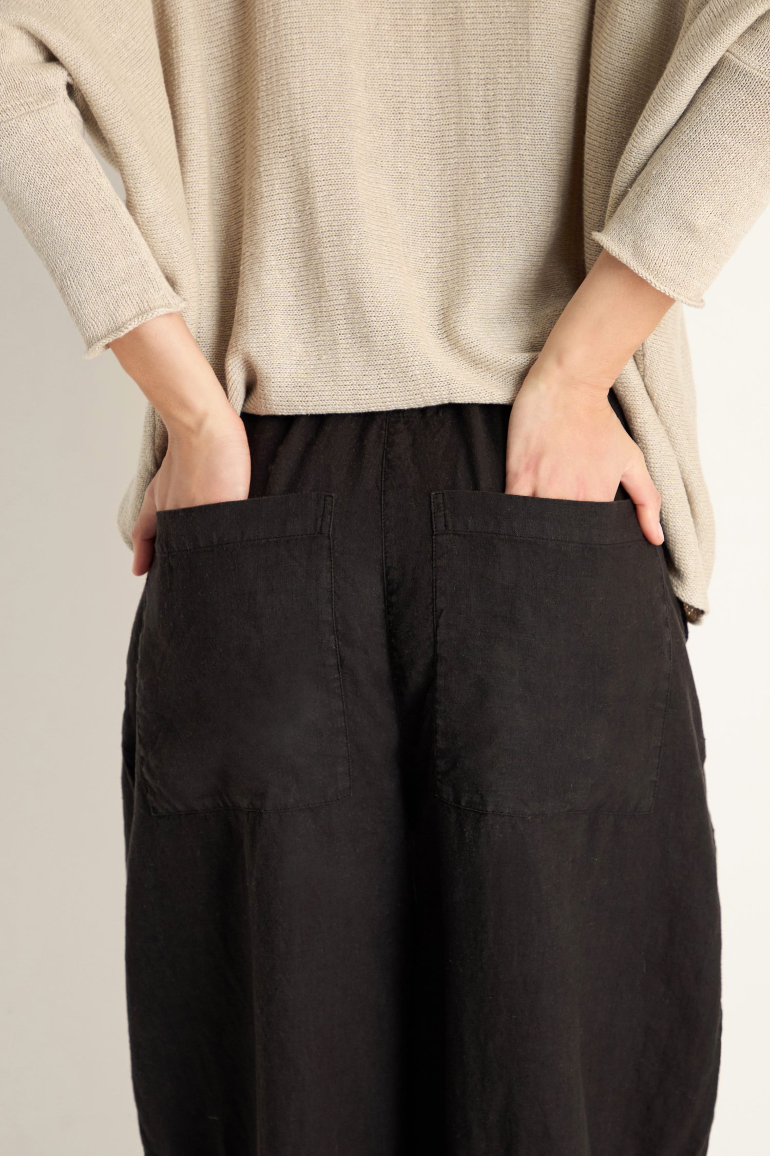 A person stands with hands in the back pockets of Curved Seam Linen Trousers in black, featuring a relaxed leg. They wear a loose beige top; only their lower back and hands are visible.