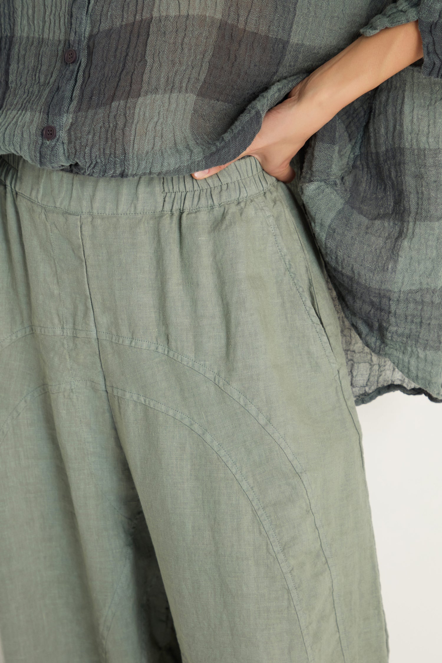Person wearing Curved Seam Linen Trousers in loose sage green with a relaxed leg and elastic waistband, paired with a textured dark green plaid shirt. Hand is placed in the right pocket of the trousers.