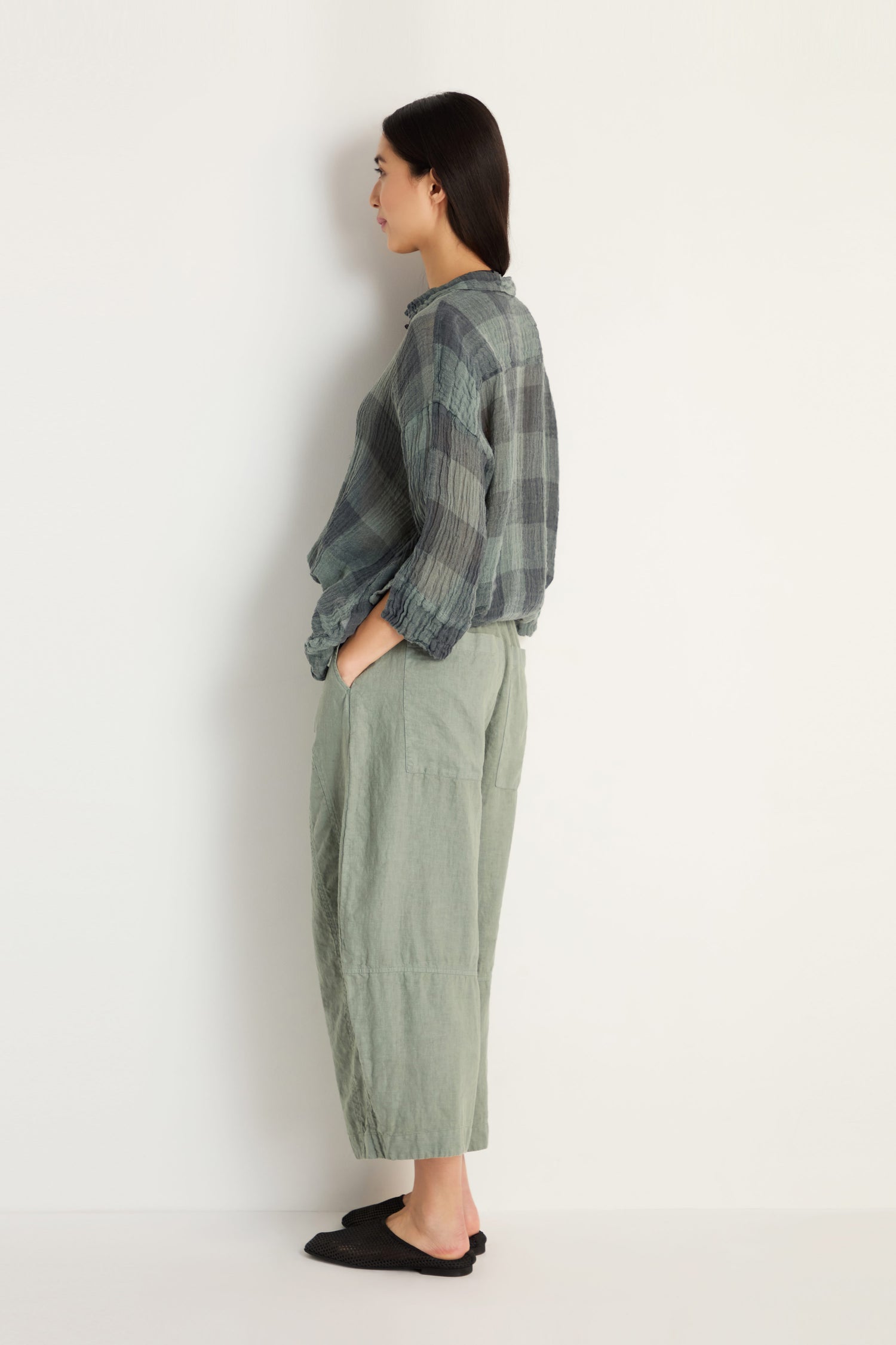 Woman in profile against a plain white wall, wearing a loose plaid shirt, black slip-on shoes, and Curved Seam Linen Trousers with relaxed legs and distinctive seam detailing.
