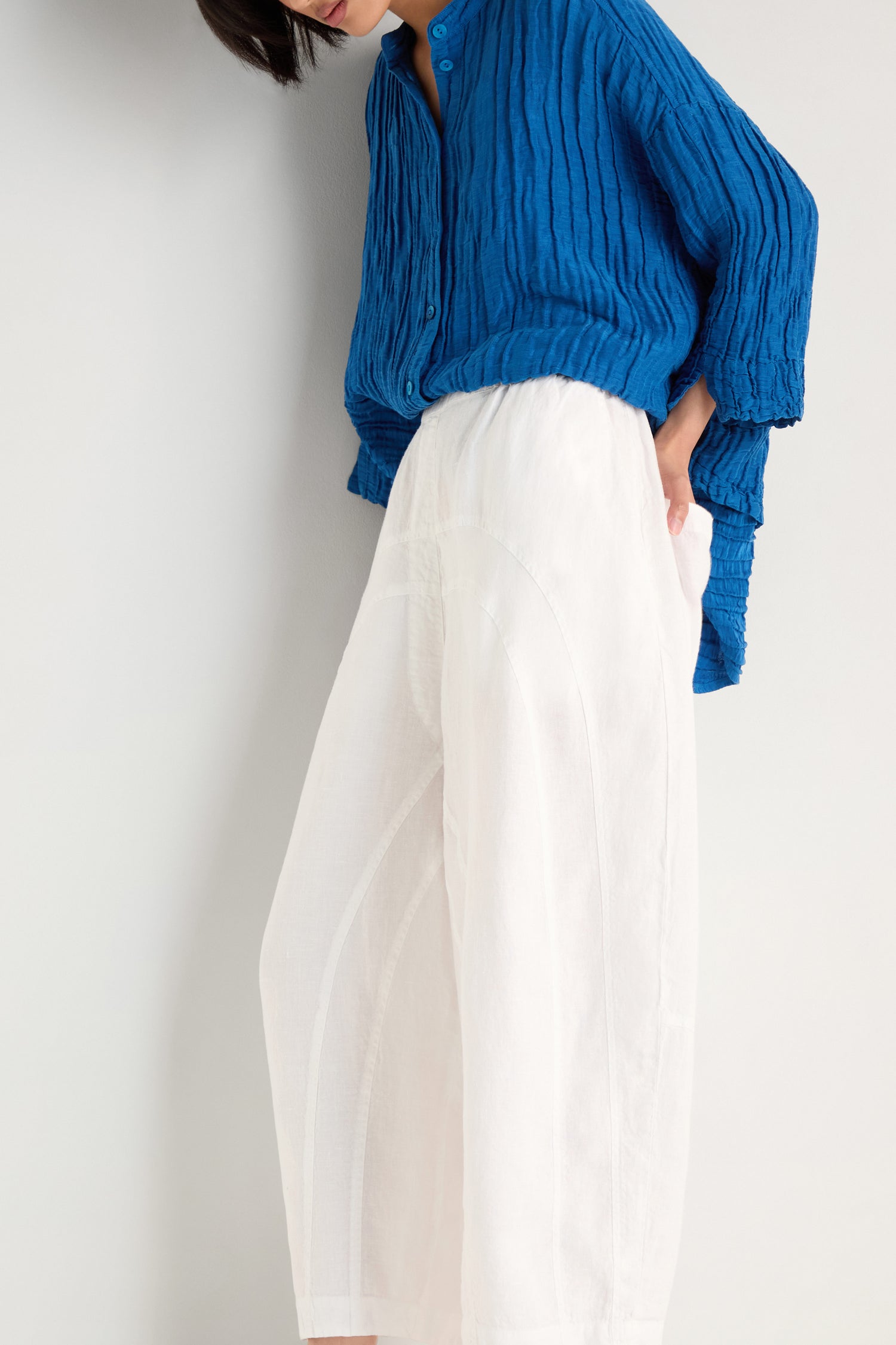 Person wearing a textured blue long-sleeve shirt and Curved Seam White Linen Trousers stands against a plain light-colored wall.