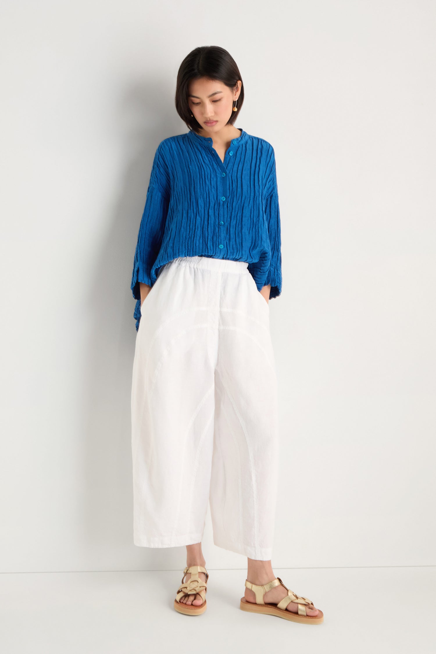 Woman stands against a white wall, hands in her pockets, wearing a blue long-sleeve blouse, Curved Seam White Linen Trousers, and beige sandals.