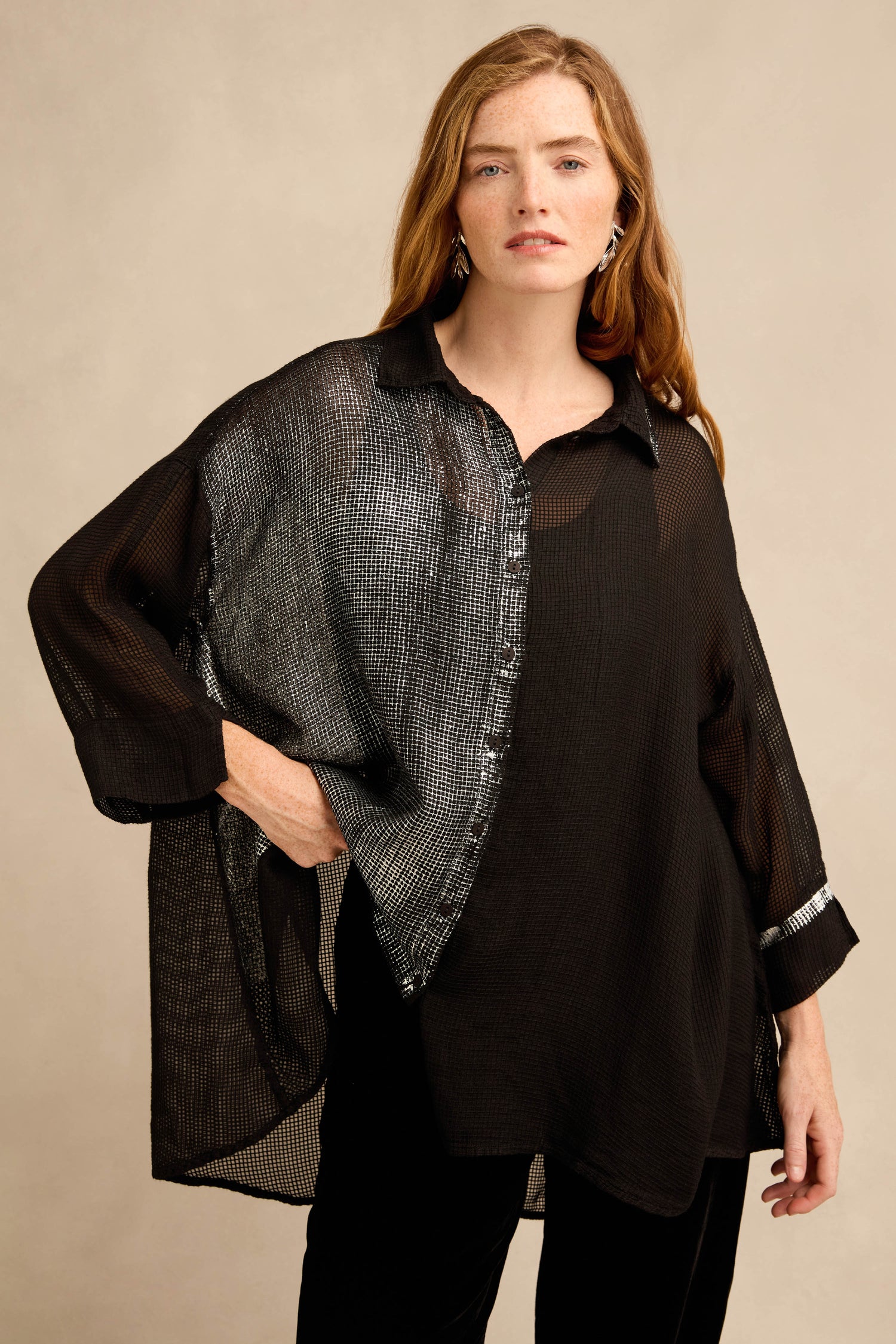 A woman stands against a neutral background wearing the Sheer Silver Panel Flared Shirt—a semi-sheer black, silk-blend top with a striking metallic silver panel—paired with black pants and silver earrings.