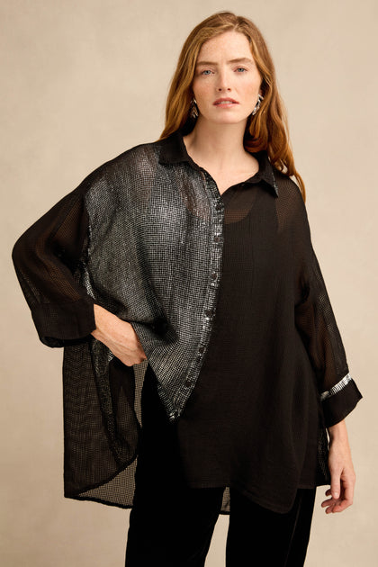 A woman stands against a neutral background wearing the Sheer Silver Panel Flared Shirt—a semi-sheer black, silk-blend top with a striking metallic silver panel—paired with black pants and silver earrings.
