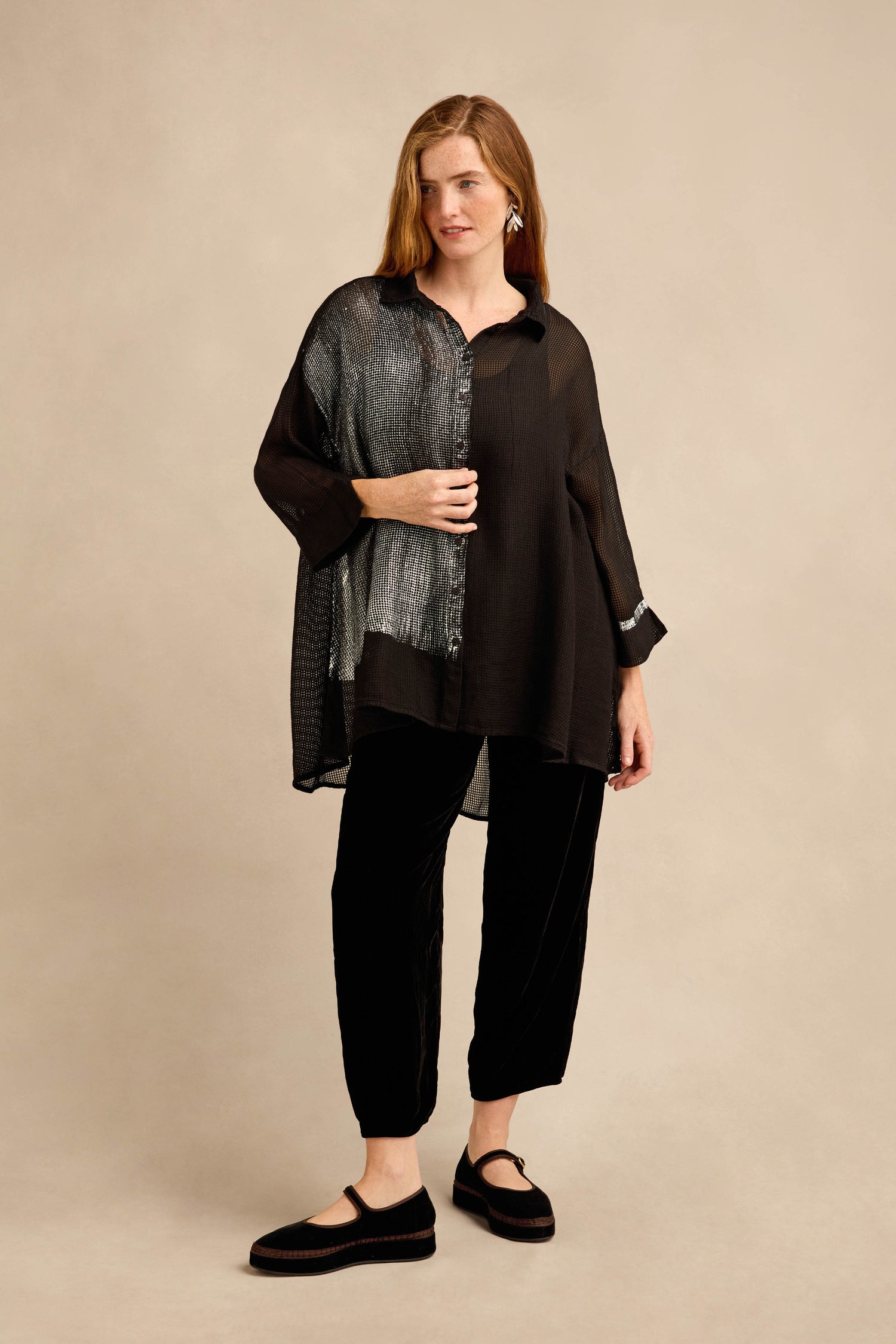 A woman stands in front of a beige background wearing the Sheer Silver Panel Flared Shirt—a loose black top with gray accents reminiscent of a metallic panel blouse—paired with black pants, black shoes, and silver earrings.